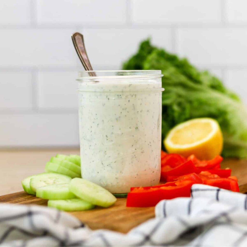 A jar of keto ranch dressing on a wooden tray with sliced raw vegetables around it.