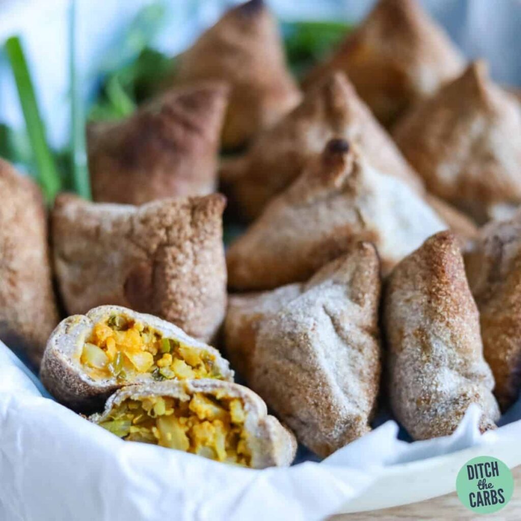 Keto samosas cooling in a dish lined with white parchment paper. One samosa has been cut in half to show the filling.