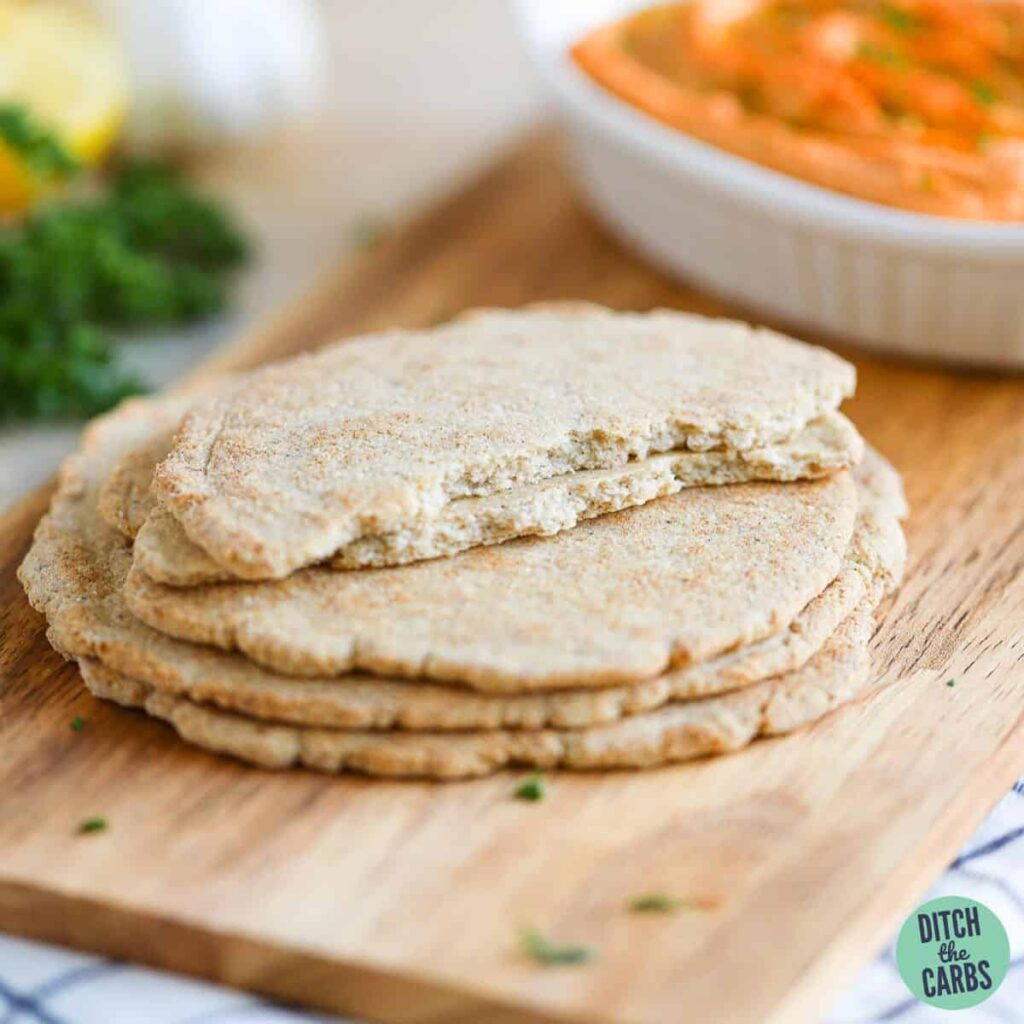 Baked low-carb pita bread stacked on a wooden tray. The top pita is ripped in half and stacked to show the texture of the bread.