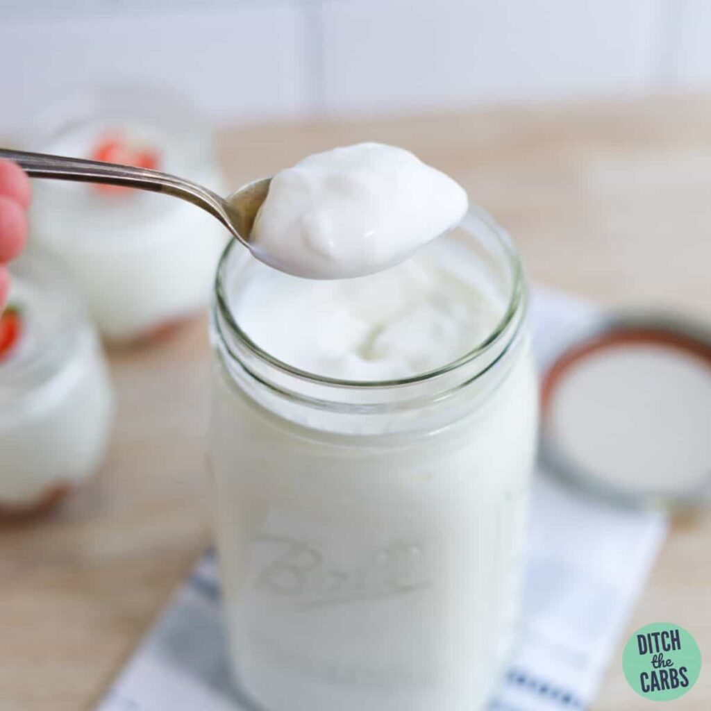 A spoon lifting a spoonful of low-carb Greek yogurt in a glass jar resting on a towel.