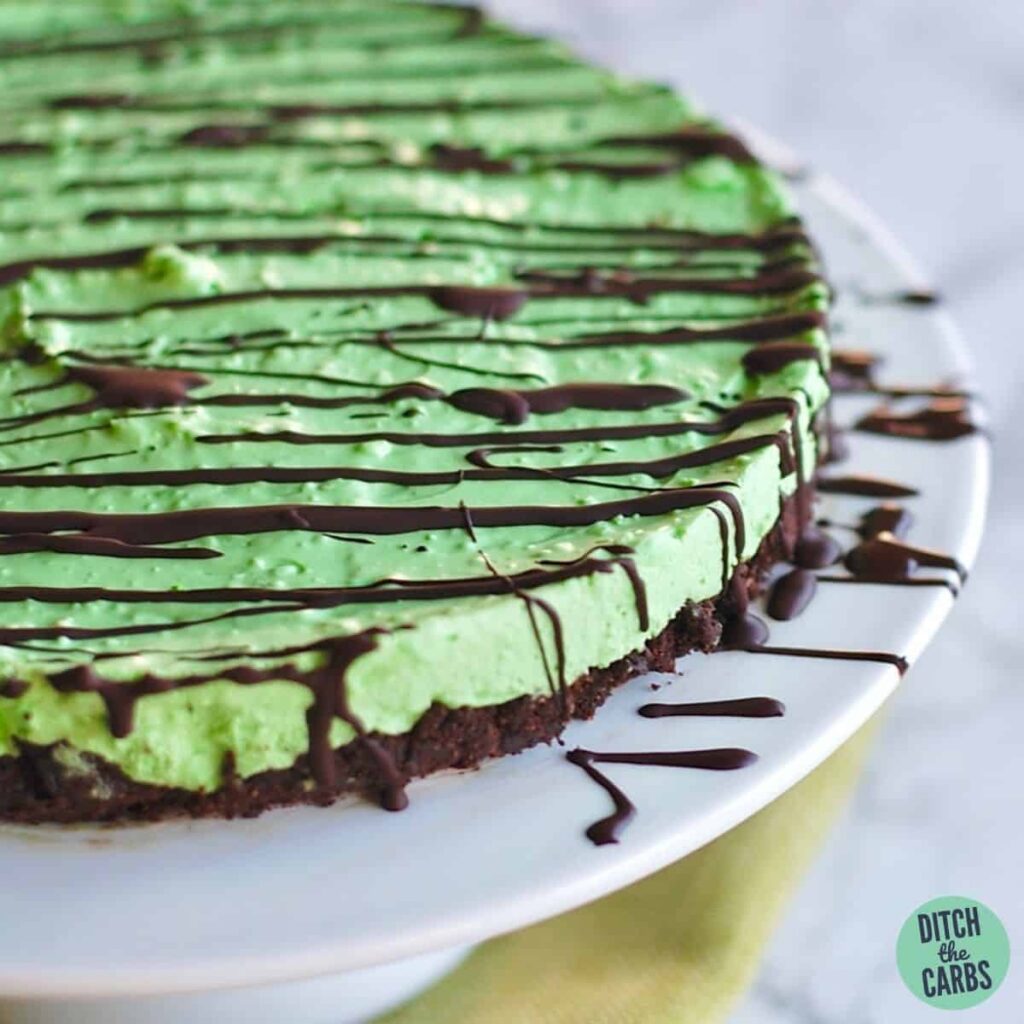 No-Bake Sugar-Free Mint Cheesecake drizzled with chocolate and served on a white cake stand